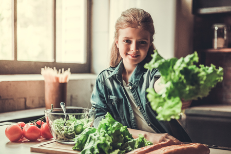 Cute teenage girl is holding lettuce, looking at camera and smiling while cooking in kitchenの写真素材