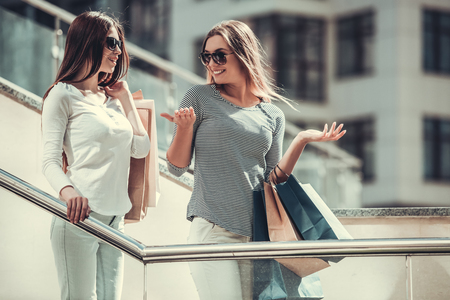Beautiful girls in sun glasses are holding shopping bags, talking and smiling while standing outdoorsの写真素材