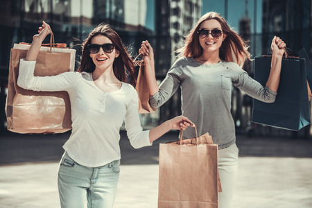 Beautiful girls in sun glasses are holding shopping bags, looking at camera and smiling while standing outdoorsの写真素材