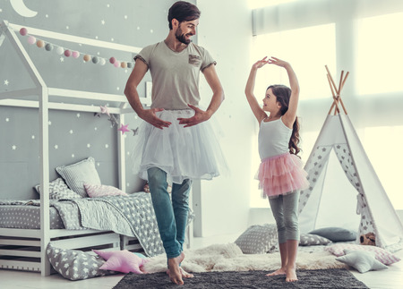 Cute little daughter and her handsome young dad in skirts are dancing and smiling while playing together in child's roomの写真素材