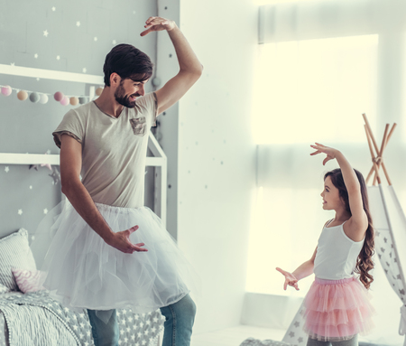 Cute little daughter and her handsome young dad in skirts are dancing and smiling while playing together in child's roomの写真素材