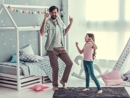 Cute little daughter and her handsome young dad are dancing and smiling while playing together in child's roomの写真素材