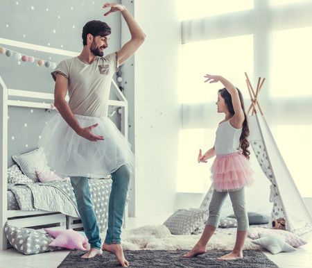 Cute little daughter and her handsome young dad in skirts are dancing and smiling while playing together in child's roomの写真素材