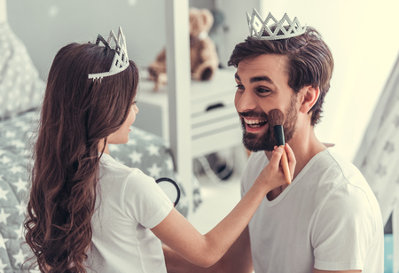 Cute little daughter and her handsome young dad in crowns are playing together in child's room. Girl is doing her dad a makeupの写真素材