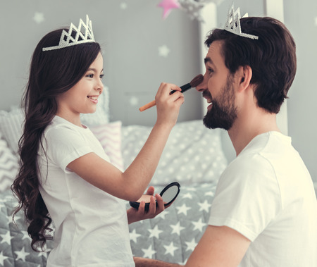 Cute little daughter and her handsome young dad in crowns are playing together in child's room. Girl is doing her dad a makeupの写真素材