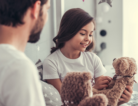Cute little girl and her handsome young dad are holding teddy bears and smiling while playing together in child's roomの写真素材