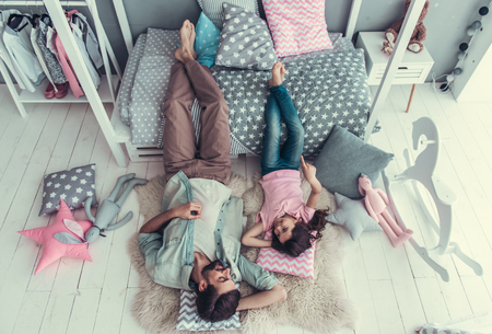 Top view of cute little daughter and her handsome young dad lying on the floor and talking while playing together in child's roomの写真素材