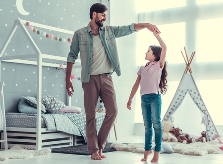 Cute little daughter and her handsome young dad are dancing and smiling while playing together in child's roomの写真素材