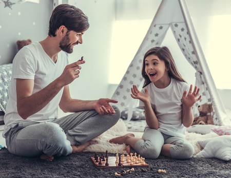 Cute little daughter and her handsome young dad are playing chess together in child's room and smilingの写真素材
