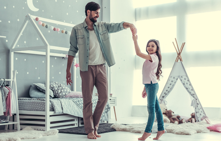 Cute little daughter and her handsome young dad are dancing and smiling while playing together in child's roomの写真素材
