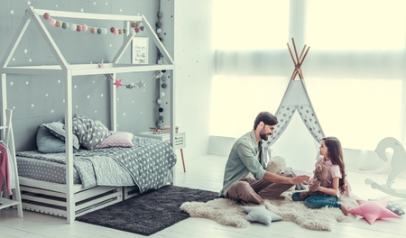 Cute little daughter and her handsome young dad are talking and smiling while playing together in child's roomの写真素材