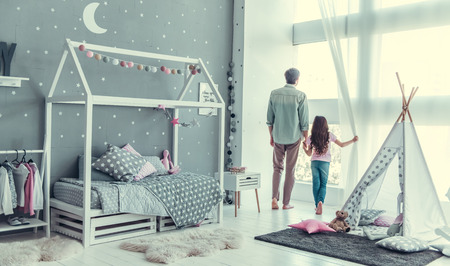 Back view of cute little daughter and her handsome young dad looking out the window and talking while spending time together in childs roomの写真素材
