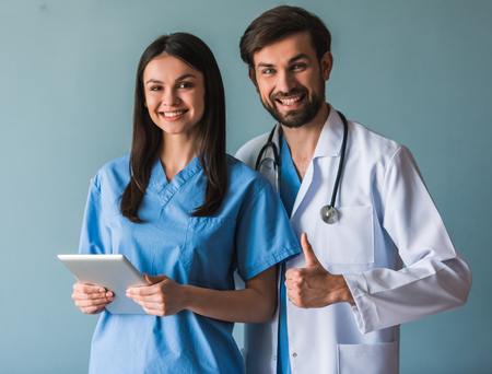 Beautiful young doctors are looking at camera and smiling, on blue background. Man is showing Ok signの写真素材