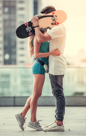 Stylish young couple is kissing behind the skateboard while walking in the city, full lengthの写真素材