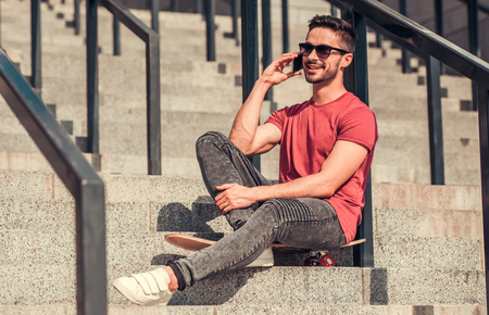 Stylish guy in sun glasses is talking on the mobile phone and smiling while sitting on his skateboard on stairs outdoorsの写真素材