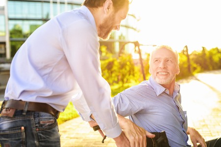 The old man is sitting in a wheelchair in the park. Behind him stands his son. The old man looks happily at his sonの写真素材