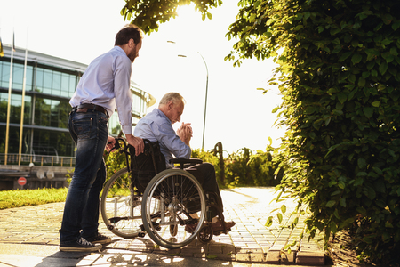 The old man is sitting in a wheelchair in the park. Behind him stands his son. Theyre walking. The old man is sadの写真素材