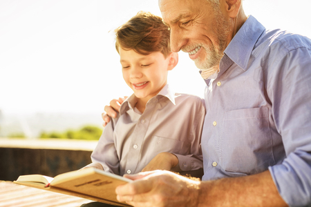 An old man shows something to his grandson in a book. They are sitting on a benchの写真素材