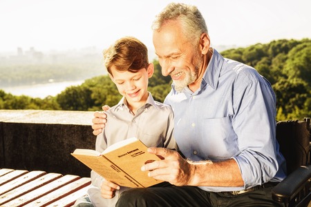 An old man shows something to his grandson in a book. They are sitting on a benchの写真素材