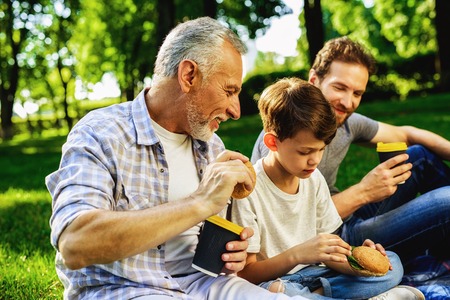 A man, his elderly father and son are sitting in a park on a picnic. A boy is sitting with a smartphone in his handsの写真素材