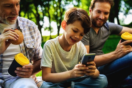 A man, his elderly father and son are sitting in a park on a picnic. A boy is sitting with a smartphone in his handsの写真素材