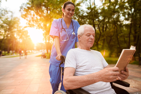 An old man who sits in a wheelchair and a nurse who is standing behind him reading a book in the park at sunsetの写真素材