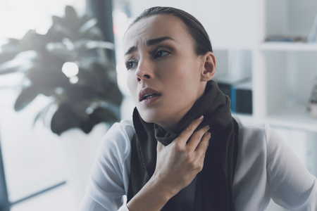 Sick woman sitting at her workplace in the office. She has a sore throat and she wore a scarfの写真素材