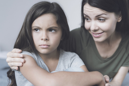 The woman embraces the girl and smiles, while the girl turns away from her and frowns sideways