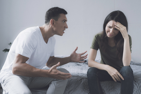 A man and a woman quarrel, sitting on the edge of a gray bed in his bedroom. A man screams at a womanの写真素材