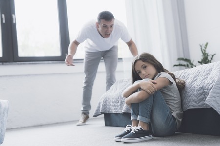 The man screams at the girl who sits on the floor near the bed with her arms around her legs and looks sadly asideの写真素材