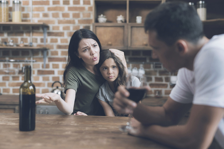 A man is sitting and looking at his wife, who is hugging their daughter and reproachfully looks at himの写真素材