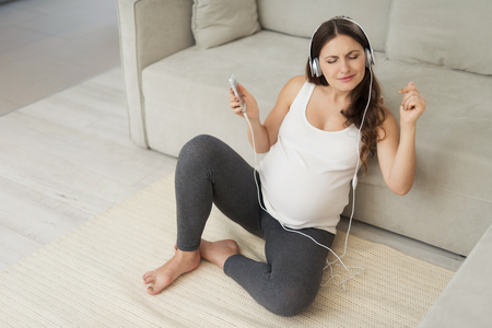 A pregnant woman sits on a light floor at home. She wore headphones and listened to music by...
