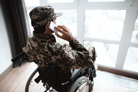The disabled in military uniform sits in a wheelchair at a large panoramic window. He rastshgovarivaet with someone on the phone. He is in his large modern apartment.の写真素材