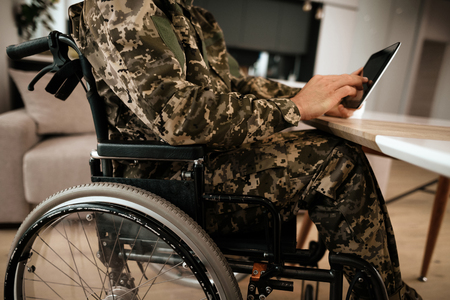 The disabled in military uniform, reading something on his modern tablet. Behind it is a large panoramic window. He is in his large modern apartment.の写真素材