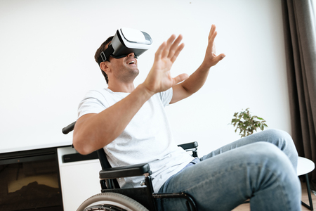 The disabled person is sitting in a wheelchair in virtual reality glasses. He spread his hands to the sides. He sits in his large bright living room and enjoys virtual reality.の写真素材