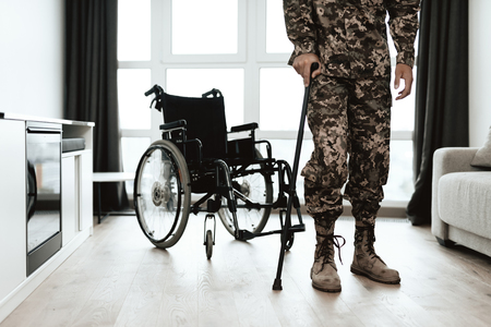 A disabled soldier is leaning on a crutch. He got up from the wheelchair and goes. Behind him is a wheelchair. He is standing in a large spacious living room.の写真素材