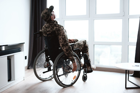 The disabled in military uniform sits in a wheelchair at a large panoramic window. He is depressed and just looks out the window. He is in his large modern apartment.の写真素材