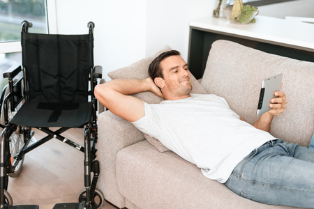 The disabled person lies on the couch and reads something on his tablet. Nearby is his wheelchair. He is on a large bright couch in his modern apartment.の写真素材
