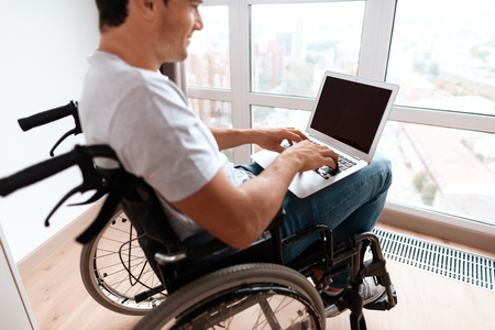 The disabled person sits in a wheelchair in front of a large panoramic window. He has a laptop on his lap, for which he works. He is in his large bright living room.の写真素材