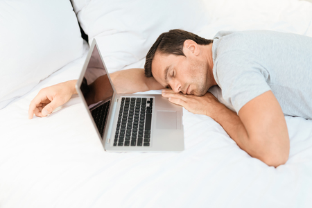 The man fell asleep on the bed before the laptop was turned on and open. He wears a gray T-shirt. He fell asleep on the big white bed.の写真素材