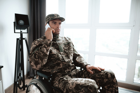 The disabled in military uniform sits in a wheelchair at a large panoramic window. He rastshgovarivaet with someone on the phone. He is in his large modern apartment.の写真素材