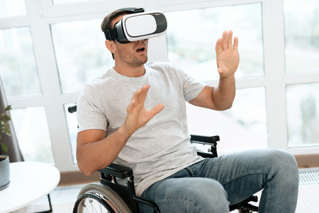 The disabled person is sitting in a wheelchair in virtual reality glasses. He spread his hands to the sides. He sits in his large bright living room and enjoys virtual reality.の写真素材