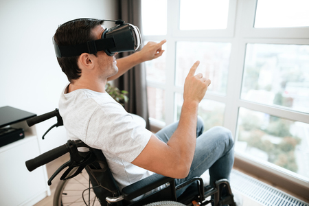 The disabled person is sitting in a wheelchair in virtual reality glasses. He spread his hands to the sides. He sits in his large bright living room and enjoys virtual reality.の写真素材