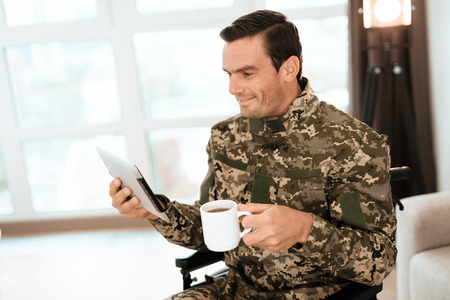 The disabled in uniform, reading something on his modern tablet and drinking coffee. Behind it is a large panoramic window. He is in his large modern apartment.の写真素材