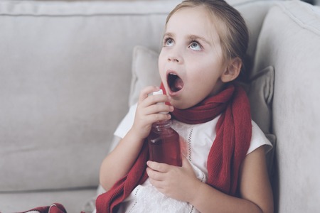 Little sick girl sits on a white couch wrapped in a red scarf. She splashes her throat with a medicinal sprayの写真素材