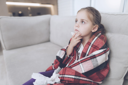 Little sick girl sits on a white couch wrapped in a red scarf. She is cold and she wrapped herself in a red blanketの写真素材