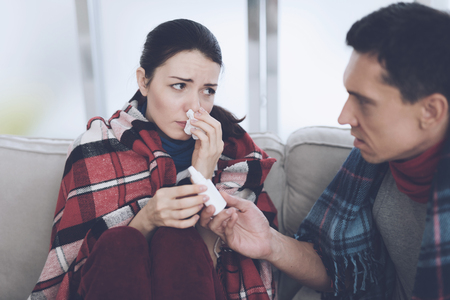 The couple is sitting on the couch wrapped in blankets. Man and woman are sick. A man gives a woman a nasal sprayの写真素材