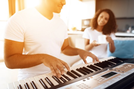 A black man sits in the living room of his apartment and plays synthesizer. A girl sitting next to him and drinking tea.の写真素材