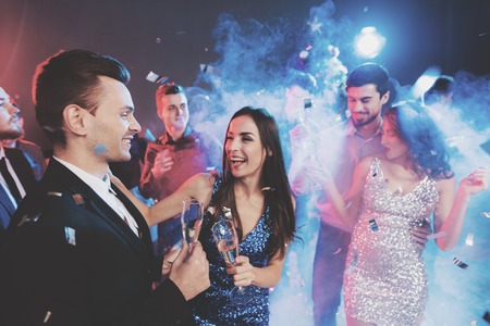 New Year party. Young couple dancing with glasses of champagne in hands. Around fly confetti and there is white smoke.の写真素材