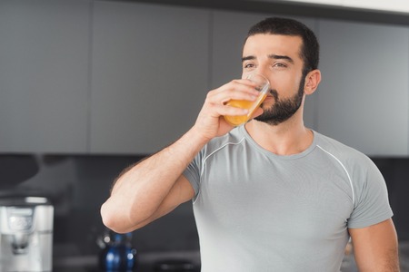 A sporty man stands in the kitchen and drinks orange juice. Next to him on a stack of vegetables and fruits.の写真素材